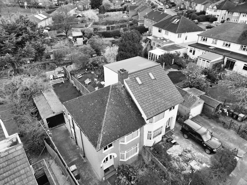 A black and white aerial photograph of a residential neighborhood with a street running vertically through the center. The image shows numerous detached and semi-detached houses with pitched roofs, so