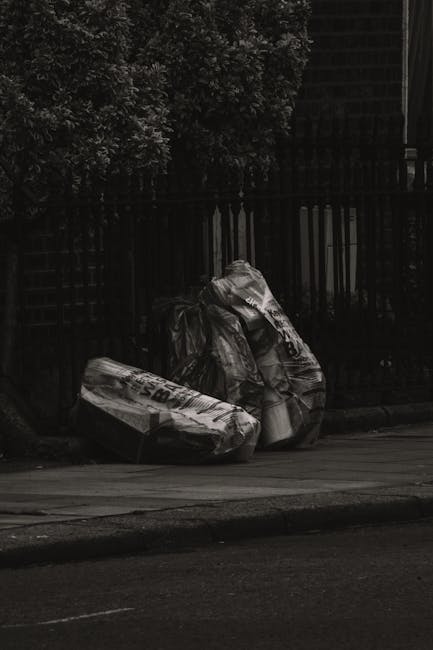 Two large black garbage bags filled with waste materials are placed on a curbside pavement next to a dark metal fence with vertical bars, behind which there are dense green foliage and trees. The bags